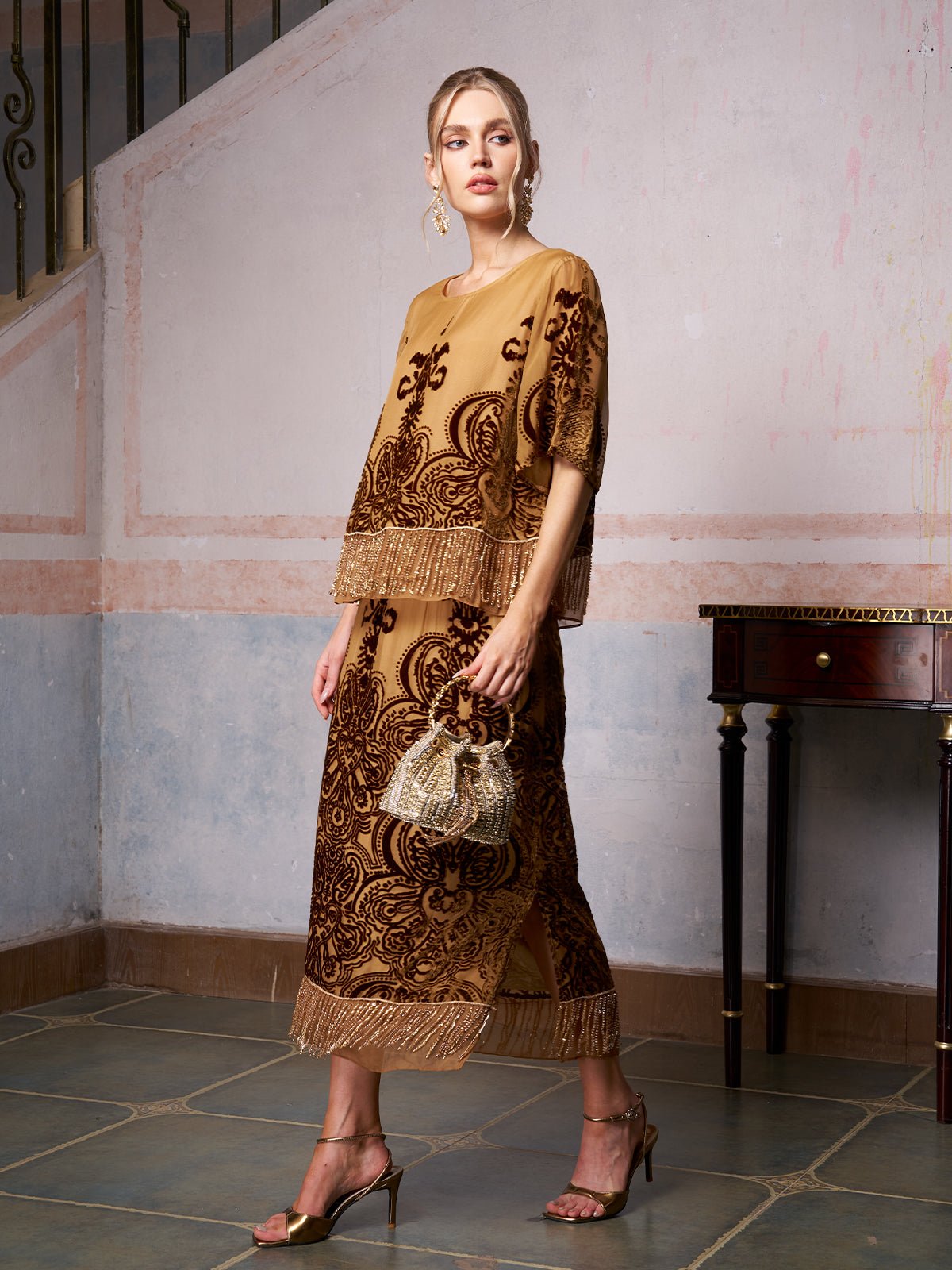 A woman stands indoors wearing the Social Queen Baroque Velvet Fringe Set with gold and brown embroidery and fringe details, paired with gold heels and a beaded handbag. A wooden table is visible in the background.