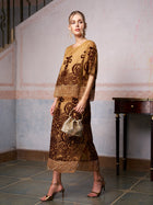 A woman stands indoors wearing the Social Queen Baroque Velvet Fringe Set with gold and brown embroidery and fringe details, paired with gold heels and a beaded handbag. A wooden table is visible in the background.