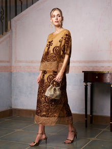 A woman stands indoors wearing the Social Queen Baroque Velvet Fringe Set with gold and brown embroidery and fringe details, paired with gold heels and a beaded handbag. A wooden table is visible in the background.