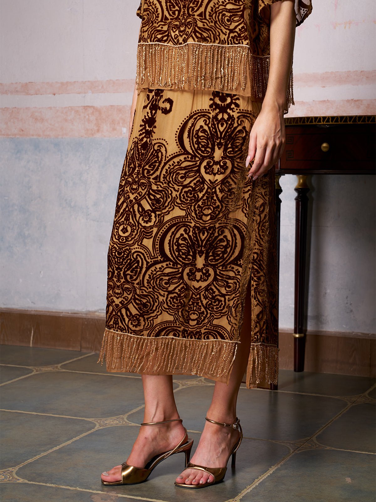 A woman in the Social Queen Baroque Velvet Fringe Set with intricate baroque patterns and fringe trim pairs it with metallic heels on a tiled floor—an elegant choice for a wedding guest.