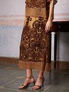 A woman in the Social Queen Baroque Velvet Fringe Set with intricate baroque patterns and fringe trim pairs it with metallic heels on a tiled floor—an elegant choice for a wedding guest.