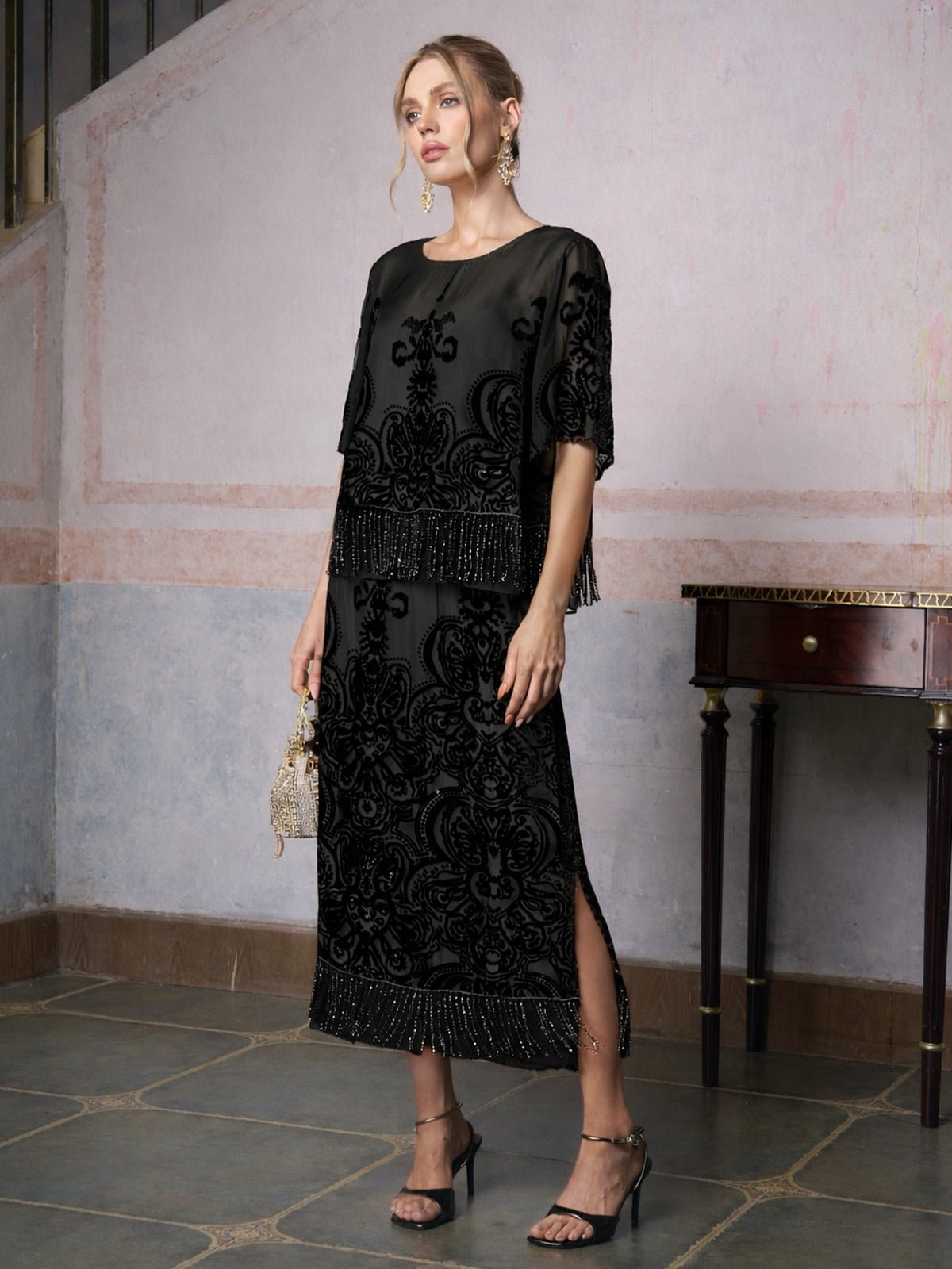 A woman wearing the Social Queen Baroque Velvet Fringe Set stands indoors by a wooden table with a small clutch—an elegant choice for a wedding guest.