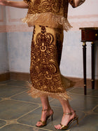 A person in the Social Queen Baroque Velvet Fringe Set, featuring gold and brown hues and fringe details, paired with gold heels, stands on tiled flooring near a small wooden table.