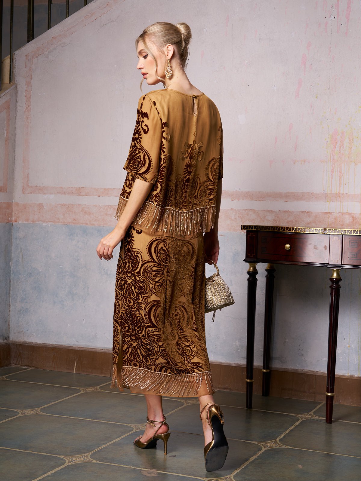 A woman stands indoors in the Social Queen Baroque Velvet Fringe Set, gold heels, and a small bag, facing away from the camera—the perfect wedding guest outfit.