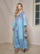 A woman with long, wavy blonde hair models the Beaded Baroque A-Line Dress by Social Queen indoors, pairing it with silver heels, a silver handbag, and jewelry.