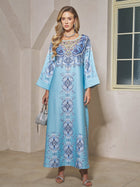 A woman stands indoors wearing the Social Queen Beaded Baroque A-Line Dress, a long light blue kaftan with baroque-inspired print and wide sleeves, accessorized with a silver handbag and sandals.