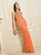A woman wears the Social Queen Beaded Crystal Strapless Gown in orange—a fitted silhouette—while standing against a cream-paneled wall, accessorized with a gold clutch and earrings.
