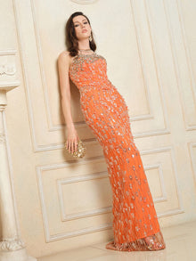 A woman wears the Social Queen Beaded Crystal Strapless Gown in orange—a fitted silhouette—while standing against a cream-paneled wall, accessorized with a gold clutch and earrings.