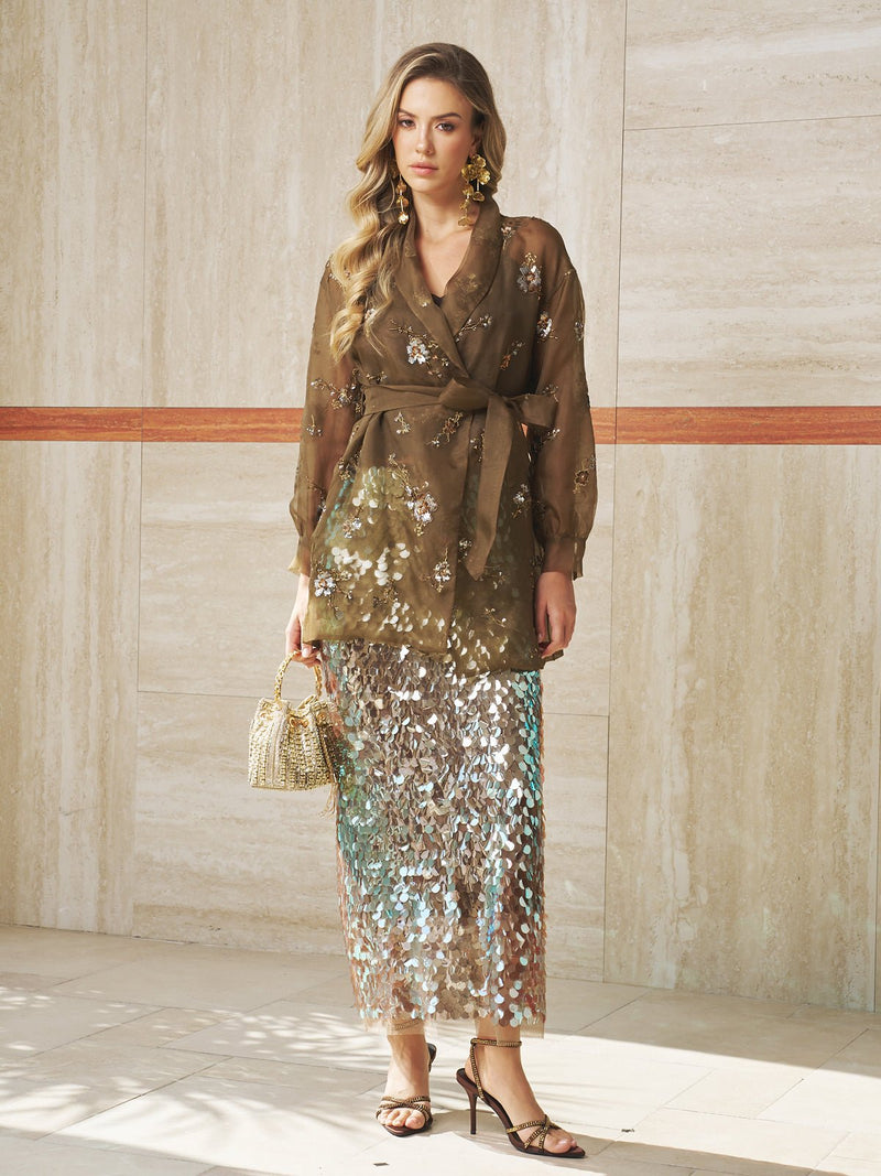 Woman stands indoors wearing the Social Queen Beaded Embroidered Silk Cardigan, paired with a shiny sequin maxi skirt, strappy heels, and holding a woven handbag.