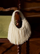 The Social Queen Beaded Fan Tassel Handbag, in cream, hangs from a chair's wooden armrest with a green cushion, its delicate beads adding charm to the scene.