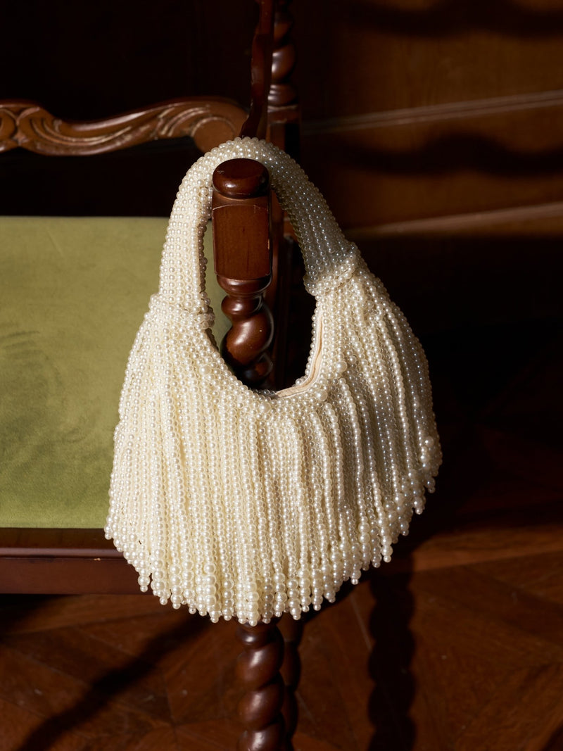 The Social Queen Beaded Fan Tassel Handbag, in cream, hangs from a chair's wooden armrest with a green cushion, its delicate beads adding charm to the scene.