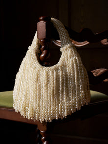 The Social Queen Beaded Fan Tassel Handbag, featuring elegant pearl strands, hangs on the carved wooden backrest of a chair with a green velvet seat.