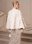 A wedding guest wears the Social Queen Beaded Floral Cape Jacket with 3D floral details and stands on stairs, holding a small purse, her hair in a bun and facing sideways.