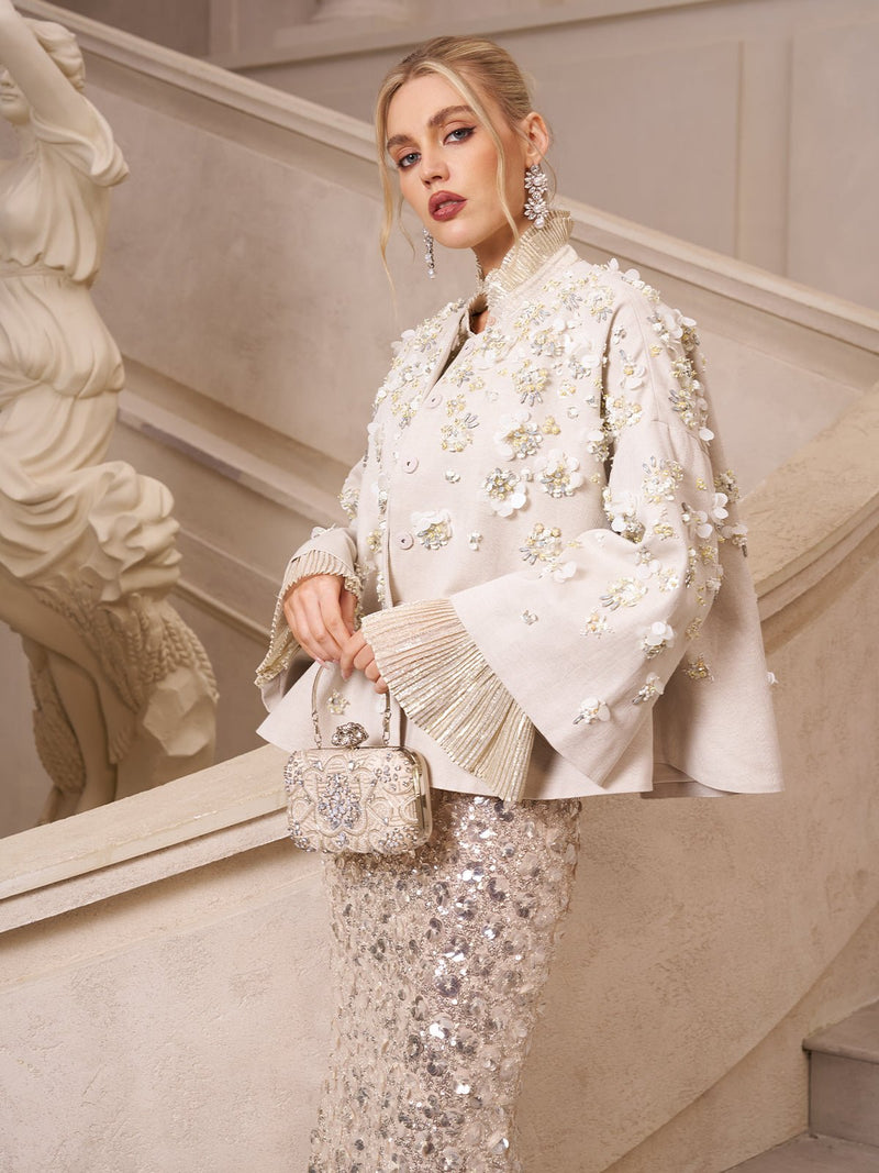 A woman stands on a staircase in the Social Queen Beaded Floral Cape Jacket, an elegant ivory piece with 3D floral embellishments, paired with a sparkling skirt and holding a small ornate handbag.