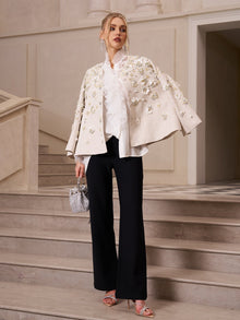 A woman stands on marble stairs in the Social Queen Beaded Floral Cape Jacket, paired with a white ruffled blouse, black pants, pink heels, and a silver mini bag—for an effortlessly stylish wedding guest look.