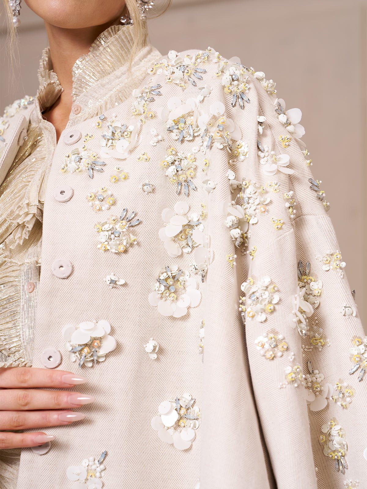 Close-up of a person wearing the Beaded Floral Cape Jacket by Social Queen, featuring cream fabric with 3D floral embellishments, sequins, and beads. The hand rests gently on this elegant wedding guest piece.