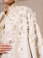 Close-up of a person wearing the Beaded Floral Cape Jacket by Social Queen, featuring cream fabric with 3D floral embellishments, sequins, and beads. The hand rests gently on this elegant wedding guest piece.