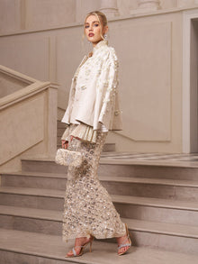 A woman stands on marble stairs wearing Social Queen’s Beaded Floral Cape Jacket, paired with heels and a matching clutch—a chic wedding guest look in an elegant, neutral interior.