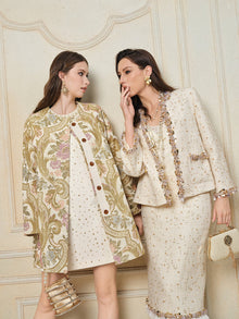 Two women in cream and gold stand together; one wears the Social Queen Beaded Floral Embroidered Tweed Jacket, leaning in to whisper to her friend. Both display statement jewelry against a neutral, decorative backdrop.