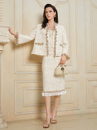 A woman poses indoors against a decorative paneled wall, wearing the Social Queen Beaded Floral Embroidered Tweed Skirt with a matching cream blazer, accessorized with a coordinating purse and strappy heels.