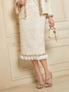 A person wearing the Social Queen Beaded Floral Embroidered Tweed Skirt with gold accents and 3D florals, paired with a small bag and gold strappy heels, stands against a beige wall.