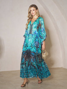 A woman in a Social Queen Beaded Floral Print Ruffled Dress with blue and turquoise beads holds a small handbag indoors, standing on beige tiled flooring against a light-colored wall.