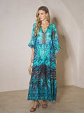 A woman wears the Social Queen Beaded Floral Print Ruffled Dress with gold accessories, holding a small woven handbag while standing indoors against a neutral wall.