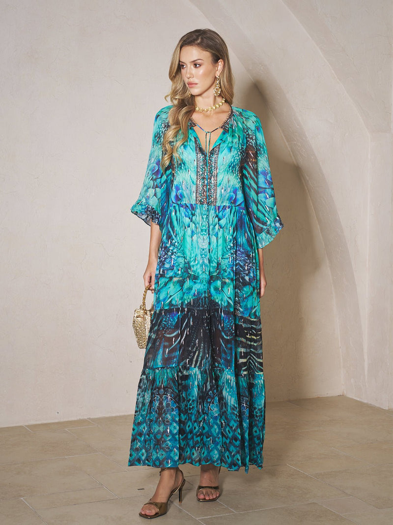 A woman stands indoors wearing the Social Queen Beaded Floral Print Ruffled Dress, paired with a small handbag, heels, and gold jewelry.