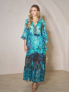 A woman stands indoors wearing the Social Queen Beaded Floral Print Ruffled Dress with a gold necklace, heeled sandals, and holding a small handbag.