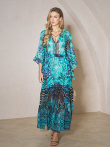 A woman stands indoors wearing the Social Queen Beaded Floral Print Ruffled Dress with a gold necklace, heeled sandals, and holding a small handbag.