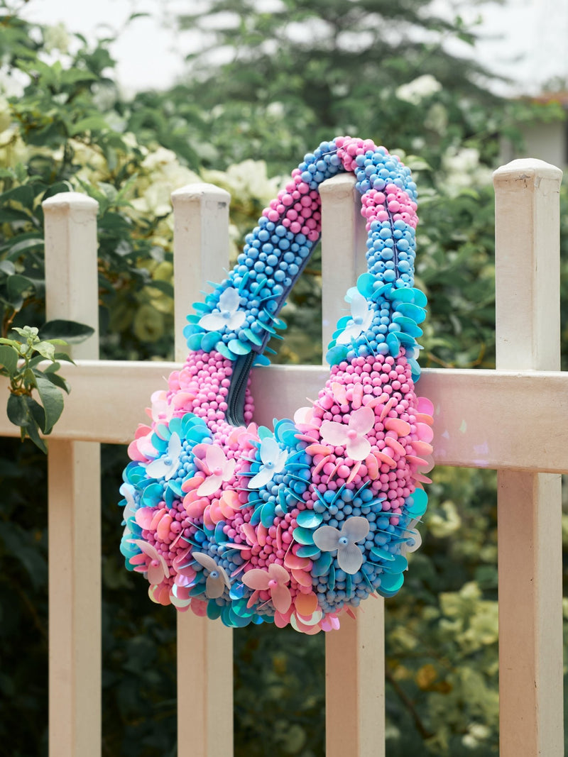 The Social Queen Beaded Floral Top-Handle Bag, featuring pink and blue hand-beading with 3D floral accents, hangs on a white fence. Its textured beads and plastic flowers pop against lush greenery in the background.