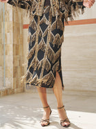 A person wears the Social Queen Beaded Fringe Midi Skirt in black and gold with fringe details, paired with gold sandals, standing on a tiled floor near a stone wall.