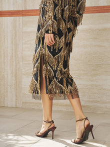A person wearing the Social Queen Beaded Fringe Midi Skirt, jazz-inspired in black and gold sequins, pairs it with brown high-heeled sandals while standing on a light tiled floor.