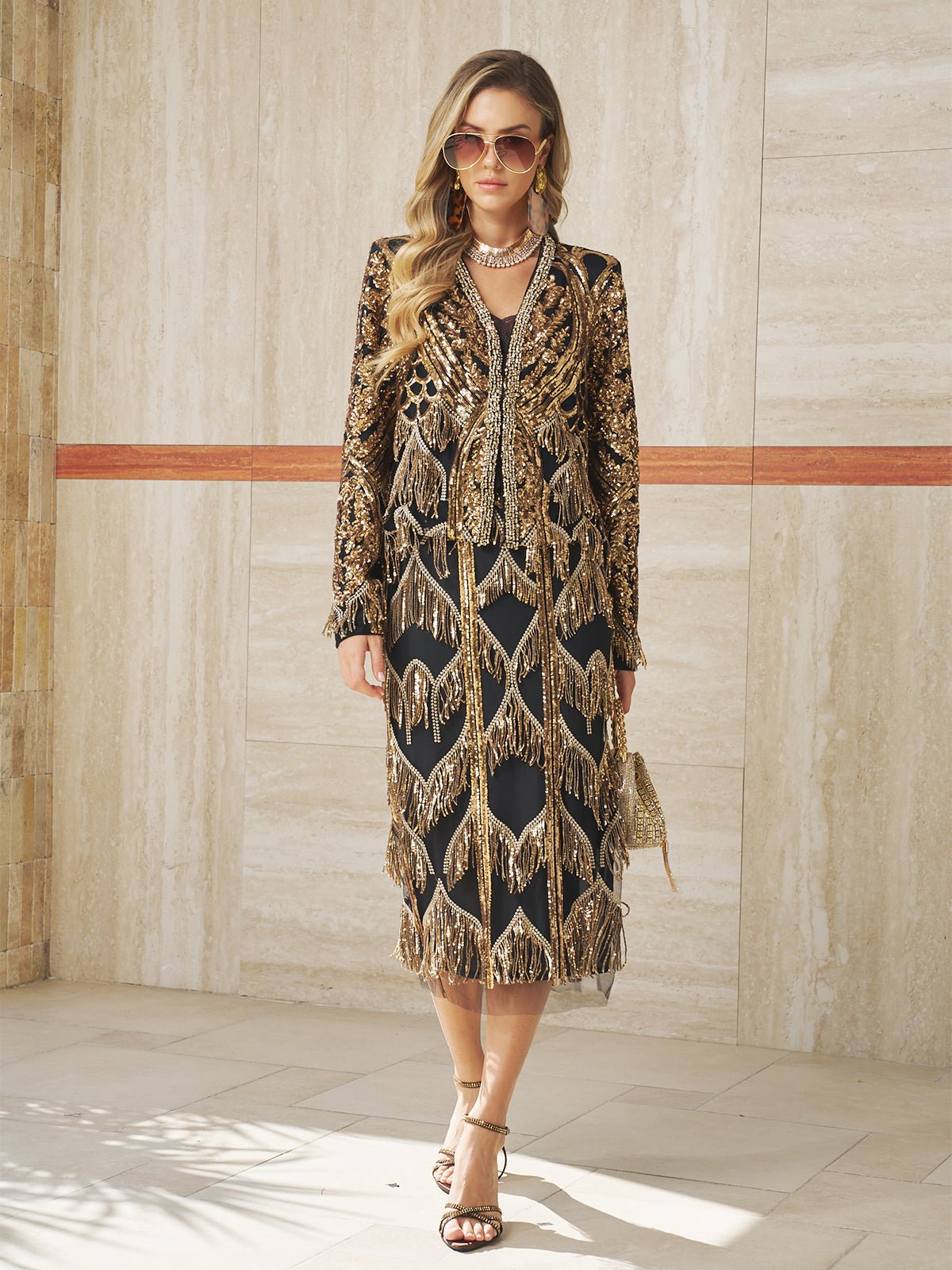 A woman wears the Social Queen Beaded Fringe Midi Skirt in black and gold, paired with sunglasses and strappy heels, standing by a light stone wall for a jazz-inspired look.