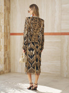 A woman stands indoors wearing the Social Queen Beaded Fringe Midi Skirt, featuring jazz-inspired black and gold embroidery with geometric fringe details. She holds a small gold handbag and wears heeled sandals.