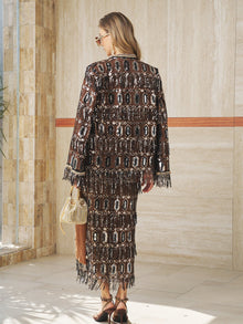 A woman stands indoors with her back to the camera, wearing Social Queen’s Beaded Fringe Sequin Midi Skirt paired with a matching jacket, heeled sandals, sunglasses, and holding a woven handbag.