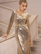 A woman in the Social Queen Beaded Gold-Accent Crystal Gown stands by a staircase, looking down with one hand on the rail—an elegant choice for wedding guests or glamorous evenings.