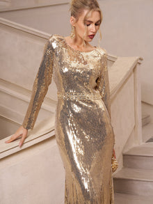 A woman in the Social Queen Beaded Gold-Accent Crystal Gown stands by a staircase, looking down with one hand on the rail—an elegant choice for wedding guests or glamorous evenings.