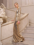 A woman stands on a staircase in the Social Queen Beaded Gold-Accent Crystal Gown, holding a matching clutch amid classical statues—a stunning wedding guest look.