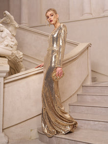 A woman stands on a staircase in the Social Queen Beaded Gold-Accent Crystal Gown, holding a matching clutch amid classical statues—a stunning wedding guest look.