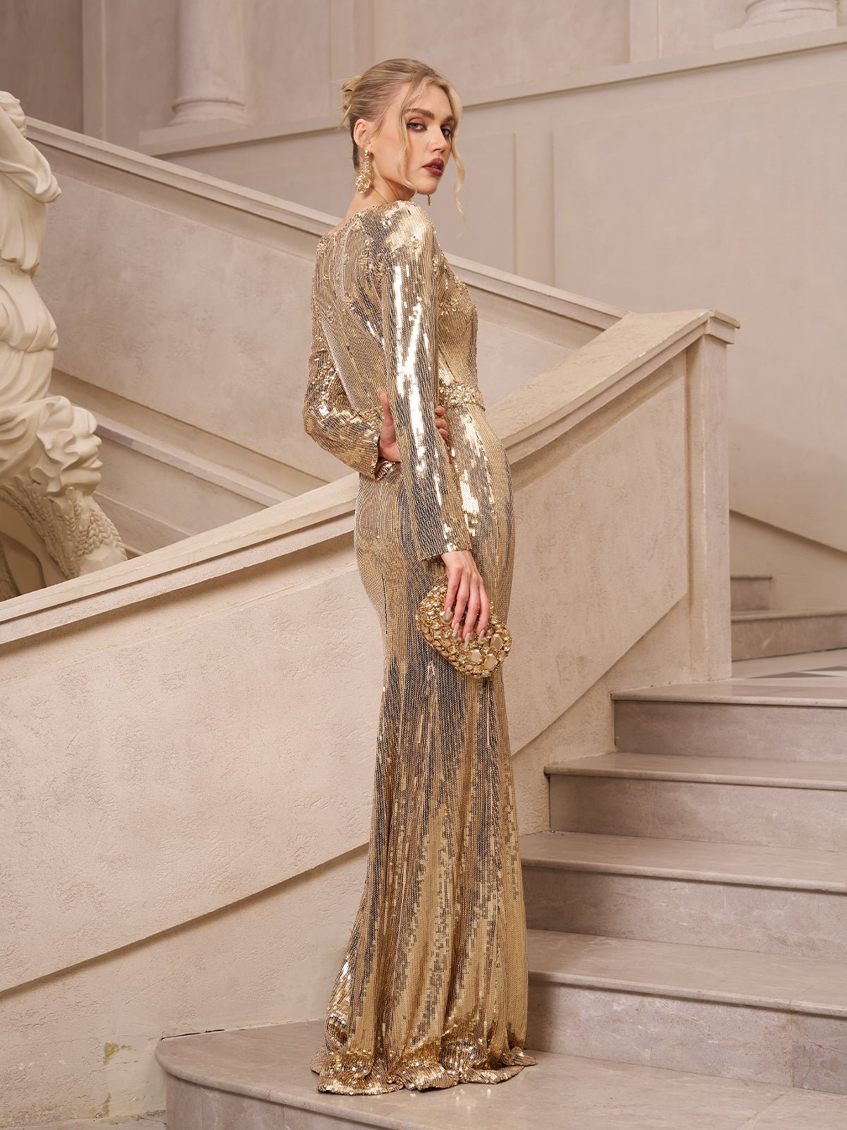 A woman in the Social Queen Beaded Gold-Accent Crystal Gown stands on a staircase, looking over her shoulder and holding a matching gold clutch—an elegant choice for evening events or as a wedding guest.