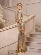 A woman in the Social Queen Beaded Gold-Accent Crystal Gown stands on a staircase, looking over her shoulder and holding a matching gold clutch—an elegant choice for evening events or as a wedding guest.