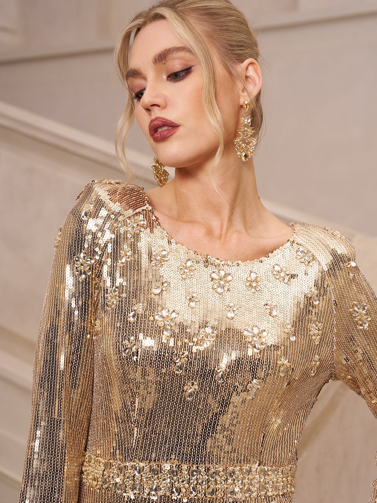 A woman in the Social Queen Beaded Gold-Accent Crystal Gown with matching earrings, ideal for a wedding guest, stands against a neutral-toned backdrop.
