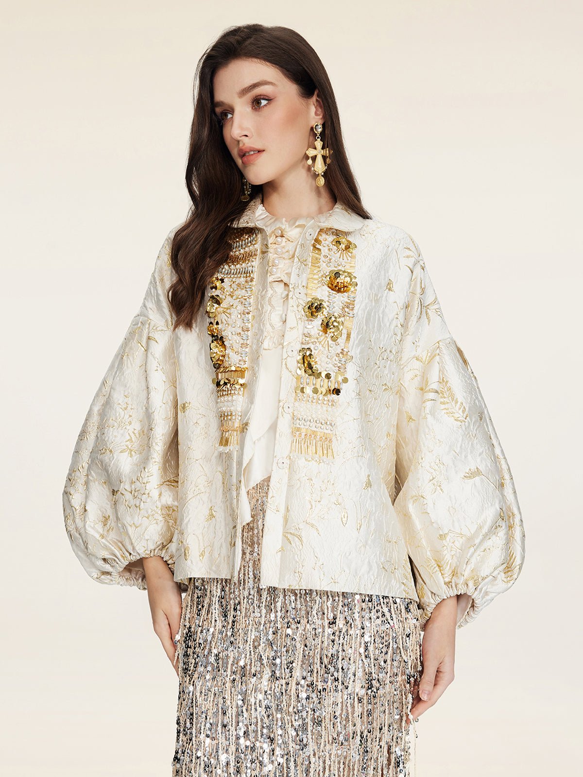 A woman wears the Beaded Jacquard Lantern Sleeve Jacket by Social Queen, featuring cream jacquard fabric with floral patterns, gold accents, and lantern sleeves, styled with a silver fringed skirt and beaded statement earrings.