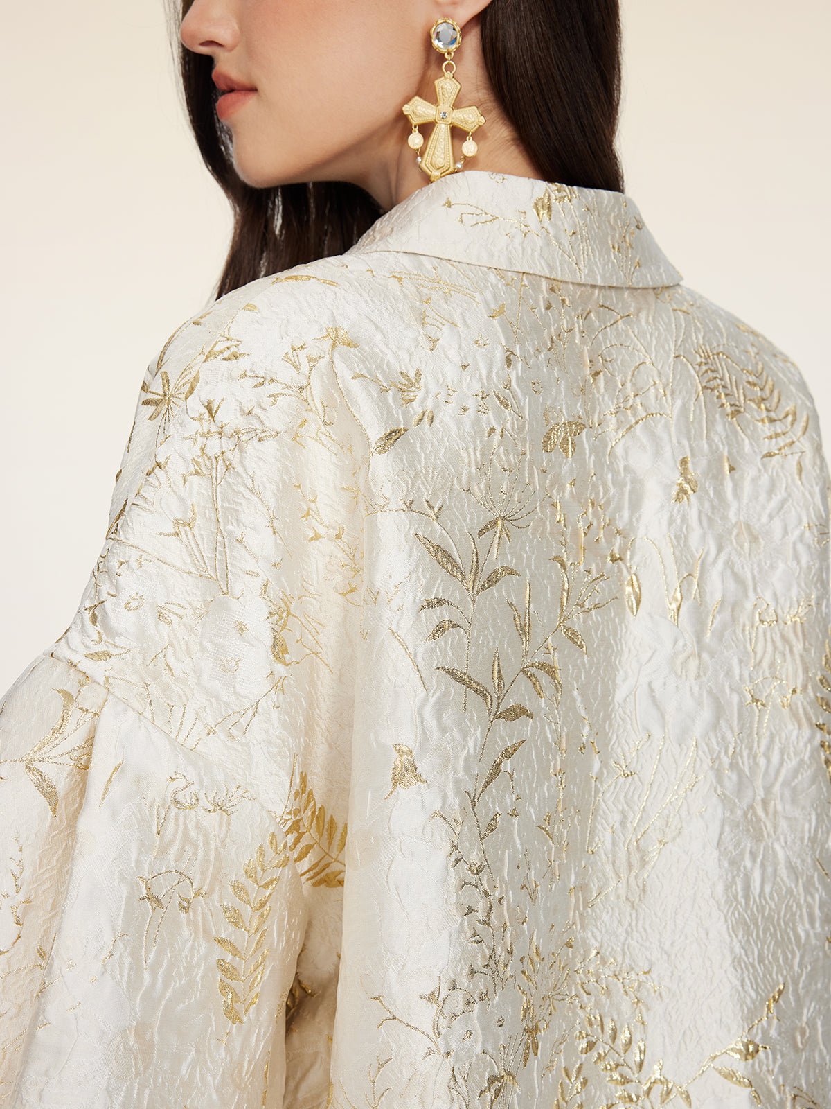 A woman shown from the side and back wears the Social Queen Beaded Jacquard Lantern Sleeve Jacket in cream and gold, paired with large gold cross-shaped earrings adorned with blue stones.