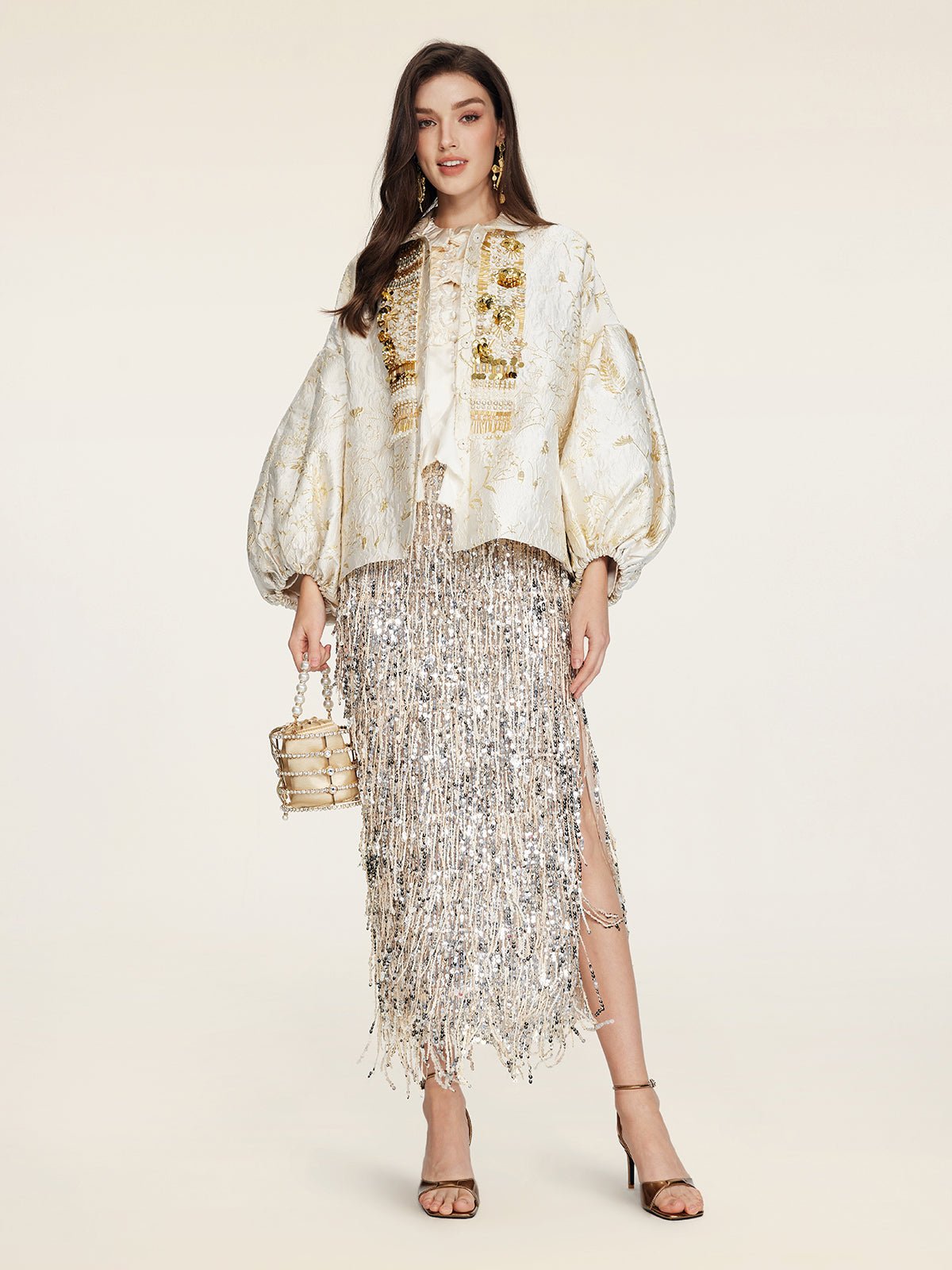 A woman wears the Social Queen Beaded Jacquard Lantern Sleeve Jacket with a silver fringed skirt, strappy heels, and a small gold handbag against a plain background.
