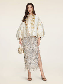 A woman wears the Social Queen Beaded Jacquard Lantern Sleeve Jacket with a silver fringed skirt, strappy heels, and a small gold handbag against a plain background.