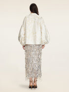 A woman faces away, wearing the Social Queen Beaded Jacquard Lantern Sleeve Jacket in cream, paired with a silver fringed hand-beaded skirt and black high-heeled sandals.