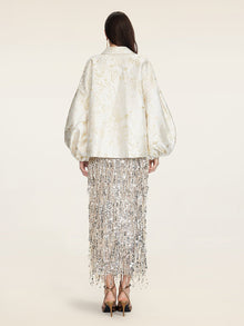 A woman faces away, wearing the Social Queen Beaded Jacquard Lantern Sleeve Jacket in cream, paired with a silver fringed hand-beaded skirt and black high-heeled sandals.