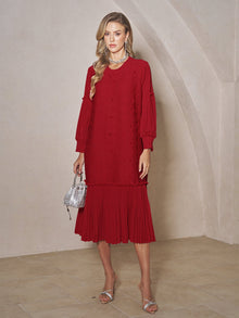 A woman stands indoors against a beige wall, wearing Social Queen's Beaded Jacquard Pleated Midi Dress in red with silver sandals, a silver necklace, and a small silver handbag.